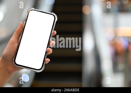 Un mockup dello schermo bianco dello smartphone è in mano a una donna sopra la scala mobile del centro commerciale sfocata sullo sfondo. Immagine ravvicinata Foto Stock