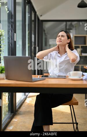 Ritratto, stanco e sovraccaricato giovane donna d'affari asiatica o operatrice femminile che soffre di dolore al collo, mal di collo, massaggiando il collo, seduta alle sue offi Foto Stock