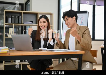 Due giovani colleghi asiatici entusiastici celebrano il successo del loro progetto aziendale insieme in ufficio, vincendo la vittoria mostrando trionfo. Foto Stock