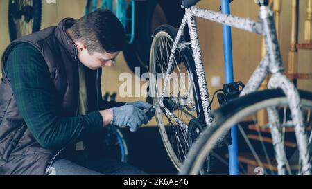 Un bel tecnico di servizio maschile nei guanti sta riparando la ruota posteriore della bicicletta utilizzando strumenti professionali. Sono visibili parti di ricambio per biciclette appese a parete e piccole officine. Foto Stock