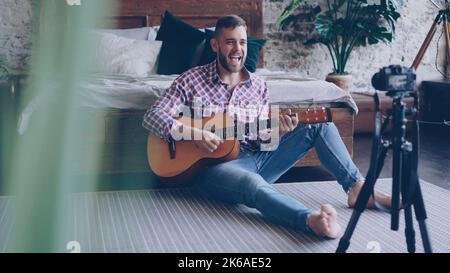 Giovane musicista bello popolare blogger sta registrando video con la macchina fotografica che suona la chitarra e canta canzone seduto sul pavimento in camera da letto. Blog e concetto di musica. Foto Stock