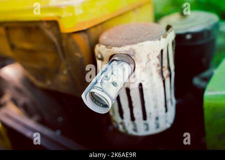 Un disegno di legge di cento dollari rotolato in un tubo sporge dal tubo di scarico di un trattore primo piano su uno sfondo sfocato. Tangenti e arresto del lavoro dovuto Foto Stock