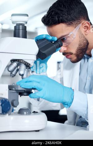 Uno scienziato di razza mista che indossa occhiali di sicurezza e guanti per l'analisi di campioni di test medici con un microscopio in laboratorio. Giovane uomo indiano che sviluppa un Foto Stock