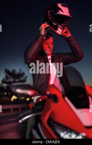 Ritratto di un uomo con una moto rossa, indossando il casco e la giacca per moto che si posano vicino alla strada al tramonto. Foto Stock
