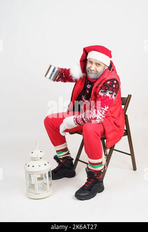 Natale e Capodanno concetto. Babbo Natale è seduto su una sedia con una tazza, c'è una lanterna nelle vicinanze. Foto Stock
