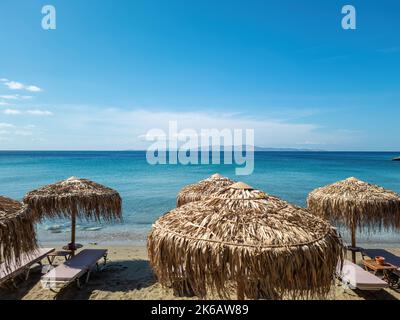Grecia. Ombrelloni di paglia lettini vuoti sotto il sole estivo greco. Spiaggia di sabbia Kalivia a Tinos isola, Cicladi. Cielo blu mare calmo sfondo. Foto Stock