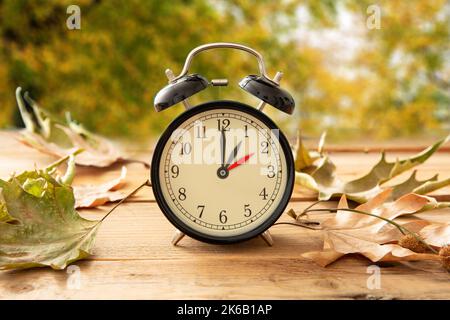 Rientro di un'ora. Ora legale, sveglia nera con cambio dell'ora su un tavolo di legno. Autunno alberi e foglie sfondo Foto Stock