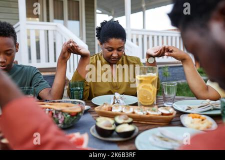 Ritratto di giovane afroamericana che dice grazia a tavola all'aperto durante la riunione della famiglia e tenendo le mani Foto Stock