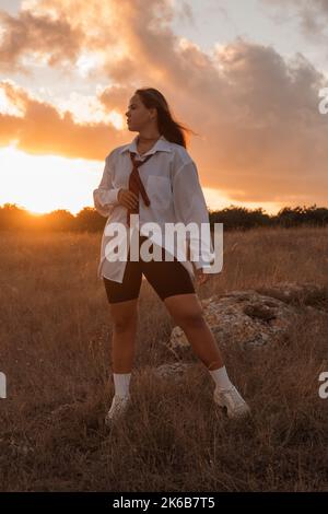 Una ragazza bianca con i capelli neri in una camicia lunga bianca, cravatta e leggings neri si alza e guarda via in un campo al tramonto. Una studentessa in un'uniforme scolastica guarda ad un futuro luminoso. Una ragazza grassa sta pensando a peso perdente. Foto di alta qualità. Foto Stock