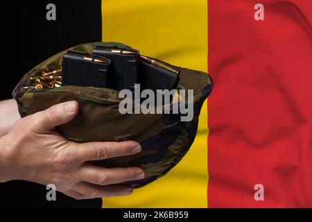 Un casco militare con cartucce e riviste per un fucile nelle mani di un uomo sullo sfondo della bandiera belga. Il concetto di Foto Stock