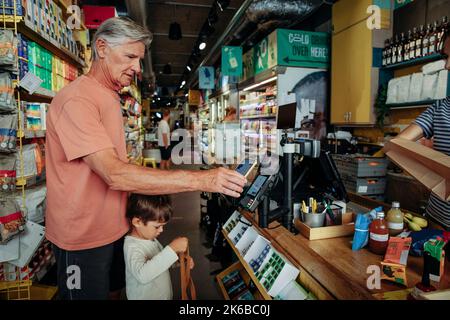 Vista laterale di un uomo anziano che effettua il pagamento online mentre si trova in piedi con il nipote al banco del check-out nel supermercato Foto Stock