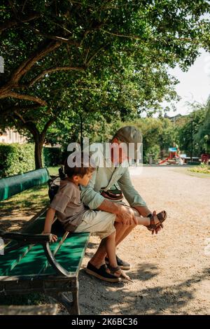 Nonno che aiuta il nipote a indossare sandalo mentre si siede sulla panca al parco Foto Stock