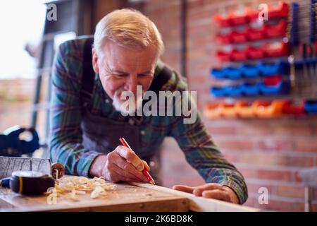 Falegname maschile senior in officina Garage misurazione e marcatura pezzo di legno con matita Foto Stock