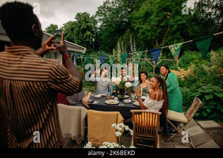 Uomo che fotografa amici di sesso maschile e femminile attraverso smartphone al party in giardino Foto Stock