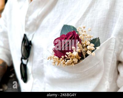 Primo piano il boutonniere in tasca di una camicia di lino bianca con occhiali da sole. Attenzione selettiva alla rosa rossa. Foto Stock