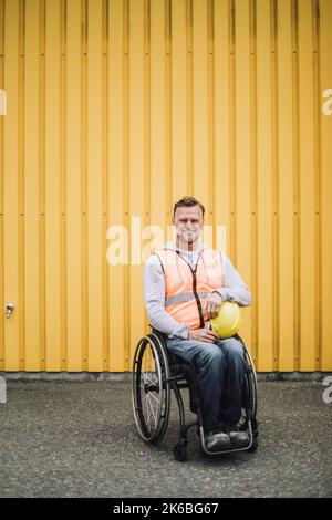 Felice lavoratore di costruzione seduto con l'elmetto in sedia a rotelle contro la parete di metallo gialla Foto Stock
