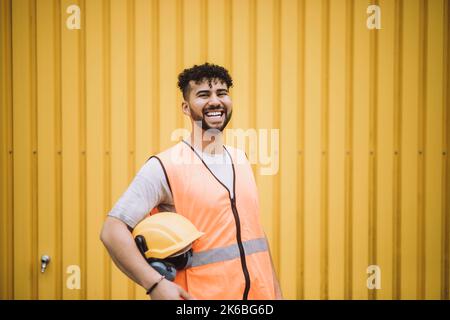 Ritratto di giovane lavoratore allegro costruzione con hardHat in piedi contro parete di metallo giallo Foto Stock