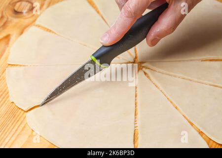 Preparazione del test per la fabbricazione di croissant francesi classici. Una donna taglia a mano con un coltello da cucina preparato pasta cruda che si trova su un tavolo di legno. Primo piano. Cottura fatta in casa Foto Stock