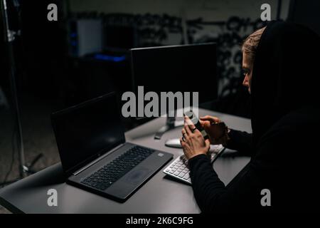 Vista dal retro dell'hacker nascosto che indossa una felpa con cappuccio impegnato nell'hacking nei sistemi di sicurezza tramite telefono cellulare Foto Stock