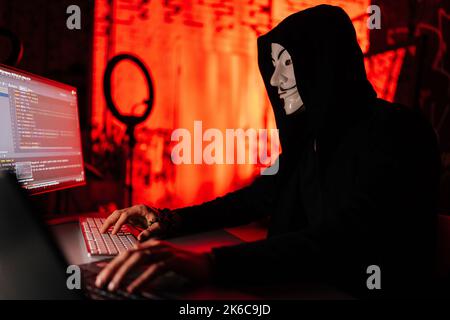 Vista laterale del ricercato hacker uomo con maschera anonima digitazione sulla tastiera wireless rompere la password utilizzando il laptop e il PC desktop Foto Stock