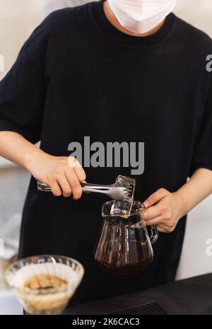 Barista con maschera per preparare caffè ghiacciato mettendo un ghiaccio in un vaso di vetro con caffè nero appena preparato. Foto Stock