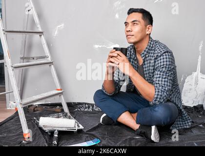 Un giovane uomo asiatico attraente siede sul pavimento prendendo una pausa del caffè dal fai da te Foto Stock