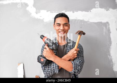 Un giovane uomo asiatico sorride mentre tiene il trapano e il martello per il miglioramento domestico del fai da te Foto Stock