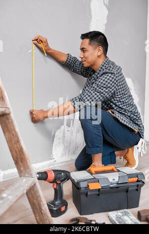 Un uomo asiatico misura contro il muro utilizzando il metro a nastro facendo casa fai da te Foto Stock