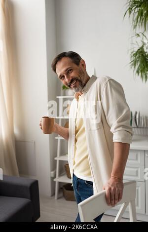 gioioso uomo bearded con tazza di tè sorridente alla macchina fotografica in cucina, immagine di scorta Foto Stock