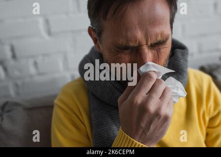 uomo malato in caldo sciarpa che tiene tovagliolo di carta e starnuti con gli occhi chiusi, immagine di scorta Foto Stock