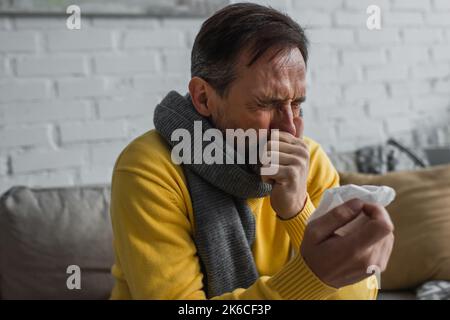 malato uomo in caldo sciarpa starnuti con gli occhi chiusi mentre tiene tovagliolo di carta, immagine di scorta Foto Stock