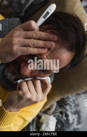 vista dall'alto dell'uomo malato con occhi chiusi che tiene tovagliolo di carta e termometro elettronico, immagine stock Foto Stock