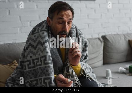 malato con gli occhi chiusi seduto sotto coperta calda e tenendo spray nasale, immagine stock Foto Stock