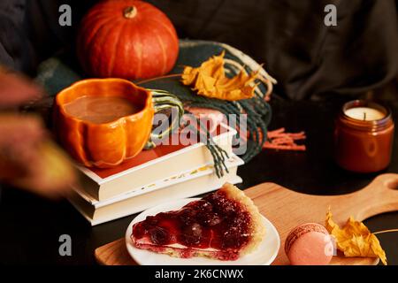 Tazza di caffè a forma di zucca, torta di ciliegie, amaretti, candela tra decorazioni autunnali. Primo piano sfocato. Casa accogliente Hygge. Foto Stock