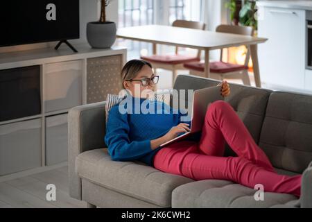 Una giovane donna sdraiata su un comodo divano lavora su un computer portatile e scrive informazioni sul business Foto Stock