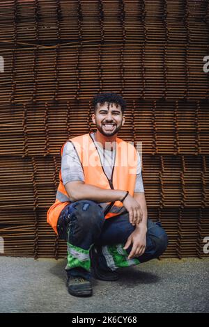 Ritratto completo di giovane lavoratore di costruzione felice accovacciato in loco Foto Stock