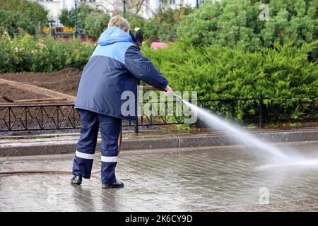 Donna operatrice in uniforme blu innaffiare il marciapiede con un tubo flessibile. Pulizia della strada e disinfezione nel parco cittadino autunnale Foto Stock