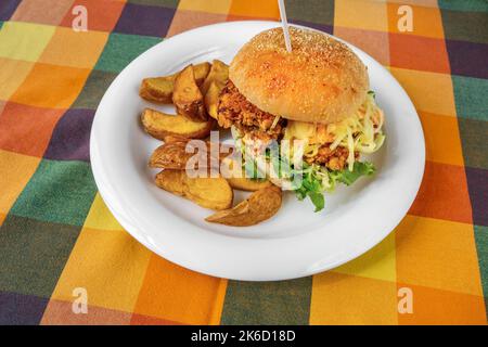 Hamburger di pollo impanato, panino con strisce di pollo, verdure e maionese, patate al forno su piatto bianco su un panno da tavolo a scacchi di colore caldo. Foto Stock