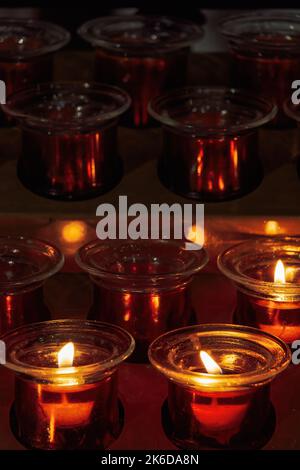 Candele bianche accese e spente di cera per preghiera votiva usate per una preghiera cristiana su un contenitore metallico con fiamme calme su uno sfondo scuro in uno stand i Foto Stock