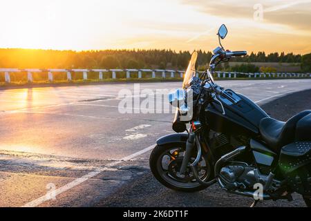 vista di una moto da crociera, classe chopper, parcheggiata da una strada asfaltata, autostrada sullo sfondo del sole al tramonto Foto Stock