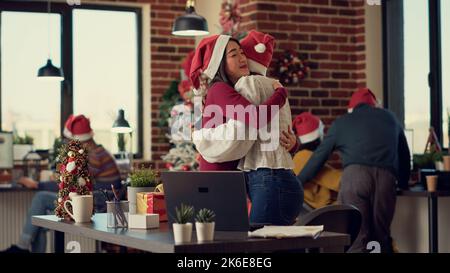 Team multietnico di colleghi che si scambiano regali di Natale durante la stagione invernale per celebrare le vacanze di natale in ufficio. Persone che danno regali sul posto di lavoro con albero e decorazioni. Foto Stock