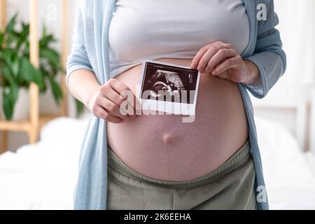 Vista dall'alto ritagliata di donna incinta con foto ecografica di feto in piedi a casa, giovane futura mamma in attesa bambino prima, gravidanza e. Foto Stock