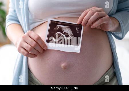 Vista dall'alto ritagliata di donna incinta con foto ecografica di feto in piedi a casa, giovane futura mamma in attesa bambino prima, gravidanza e. Foto Stock