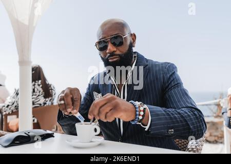Un ritratto di un ragazzo nero senza peli maturo con una barba ordinata seduta in un ristorante costiero di strada e versare lo zucchero nel suo caffè espresso c Foto Stock