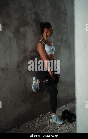 Giovane ragazza sportiva che appoggia la gamba sinistra sul muro, guardando a sinistra, palestra Foto Stock