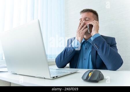 Un uomo d'affari deluso che parla con il cellulare in ufficio. Un manager in un ufficio luminoso vestito da un tuta blu parla al telefono. Foto Stock