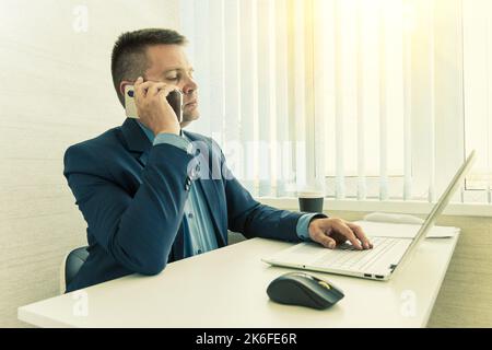 Uomo d'affari sicuro seduto alla scrivania dell'ufficio. Un manager in un ufficio luminoso vestito da un tuta blu parla al telefono. Foto Stock