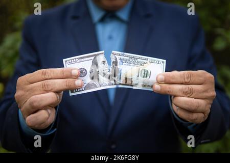 Primo piano di mani umane che strappano banconote da cento dollari. Il concetto di perdere denaro e reddito. Un uomo d'affari in una giacca strappa una fattura di carta di 10 Foto Stock