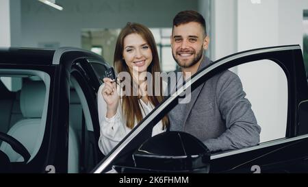 Ritratto di coppia attraente che tiene le chiavi di nuova auto dopo l'acquisto in showroom concessionaria Foto Stock