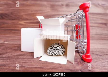 Carrello della spesa in miniatura capovolto con piccole scatole di spedizione e cervello umano su uno sfondo di legno. Cervello creativo nel concetto del supermercato. Foto Stock